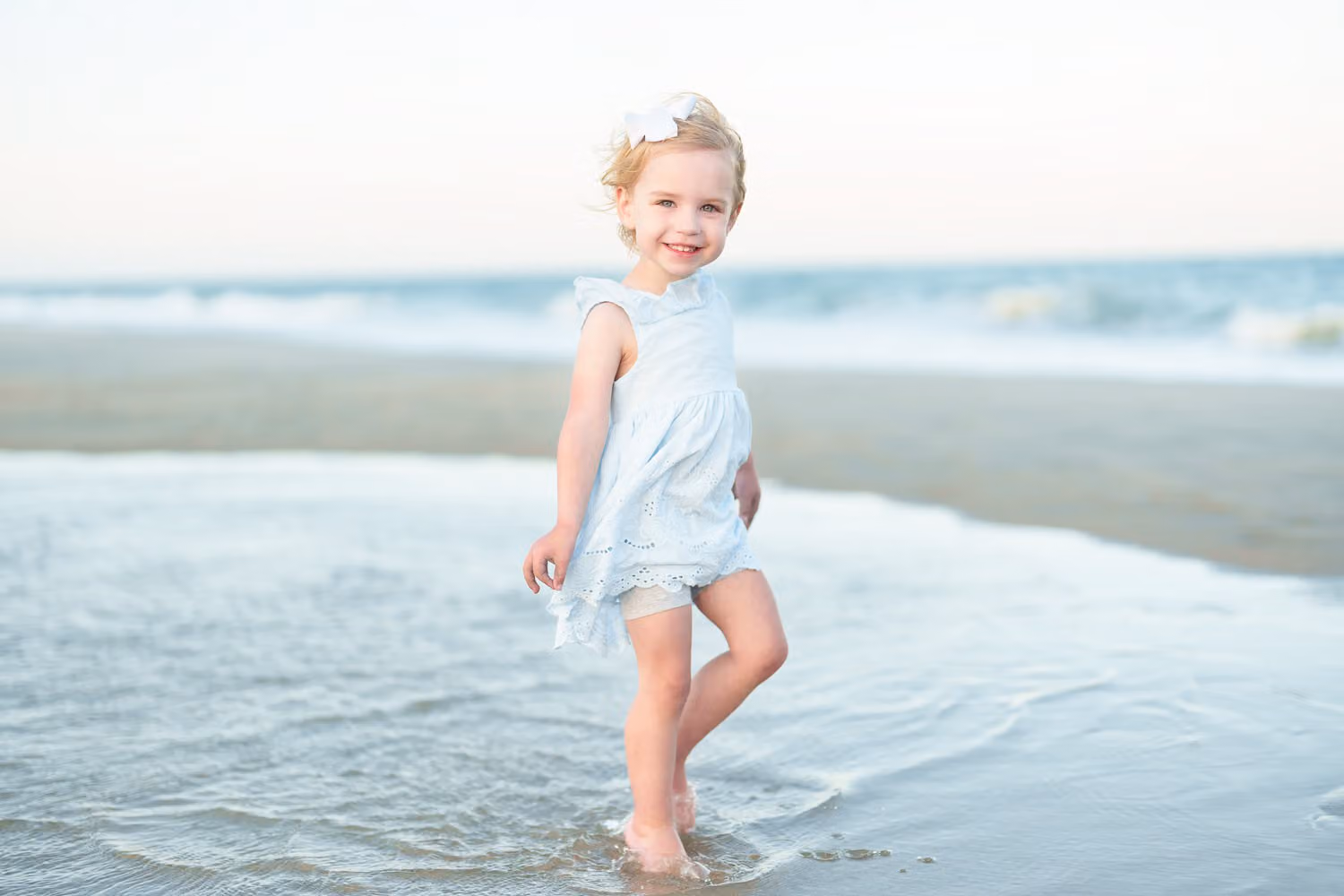 Little blonde girl in a light blue dress stepping through shallow water by the ocean at Huntington Beach State Park