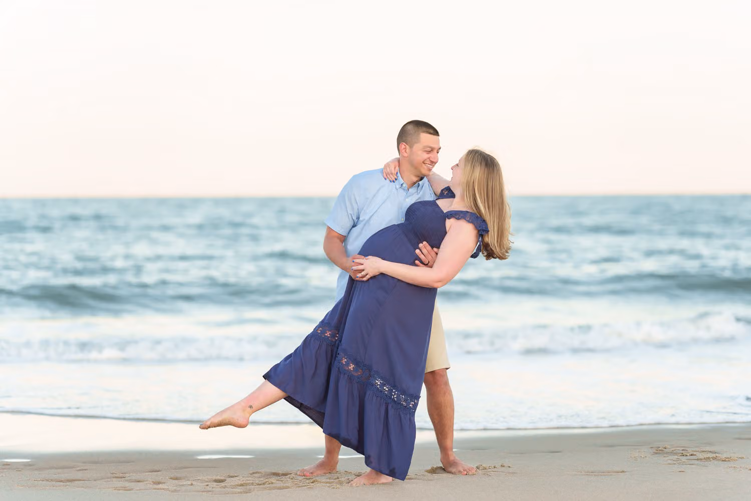 Husband dipping his expectant wife back in a navy dress in front of the ocean at Huntington Beach State Park