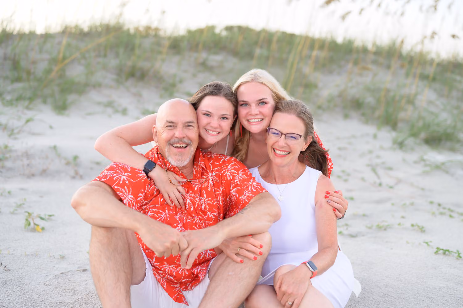 Dad in a red shirt and mom hugging their two grown daughters in the sand by the dunes at Huntington Beach State Park