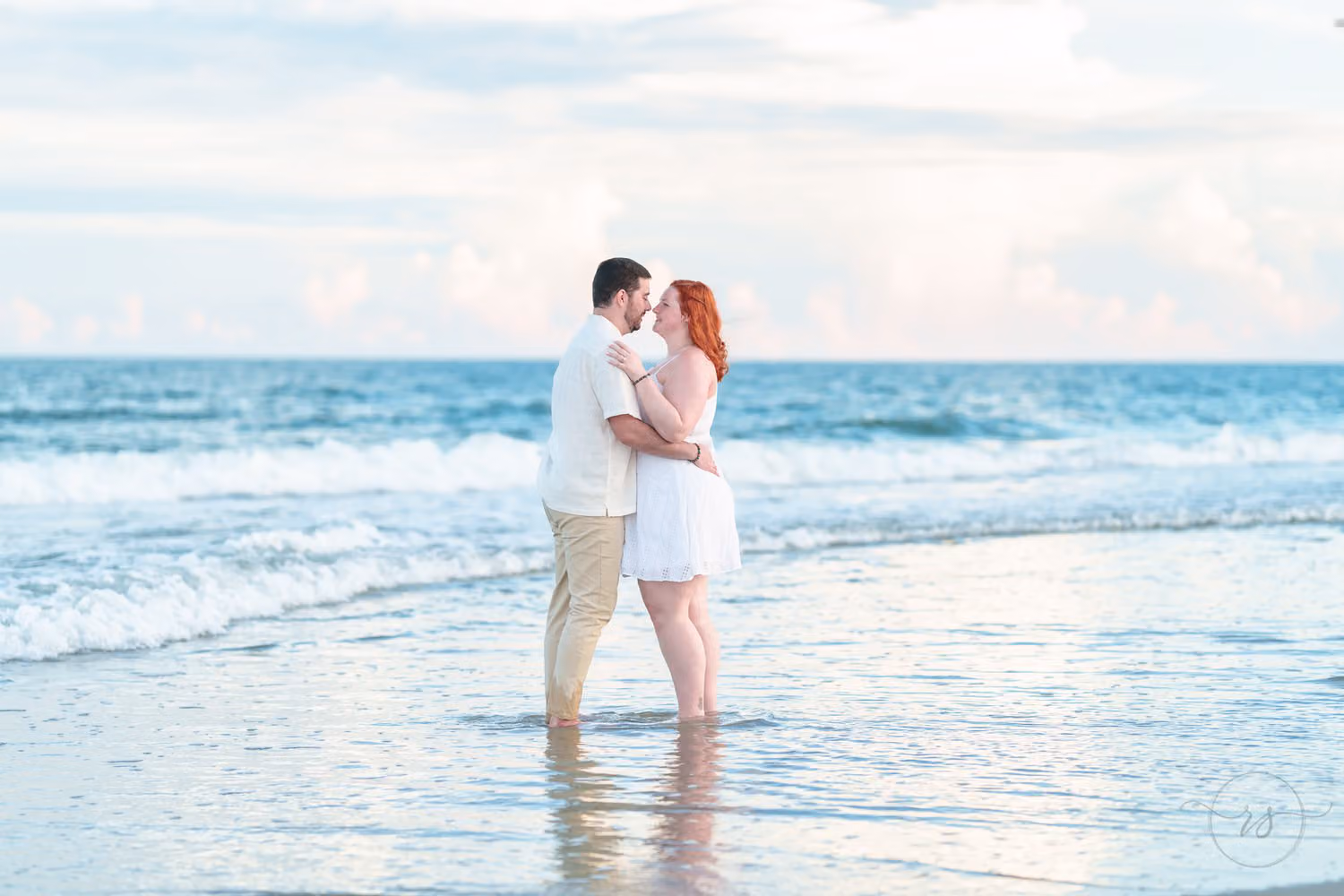 Couple standing in shallow surf with foreheads close for a kiss under pink and blue evening clouds