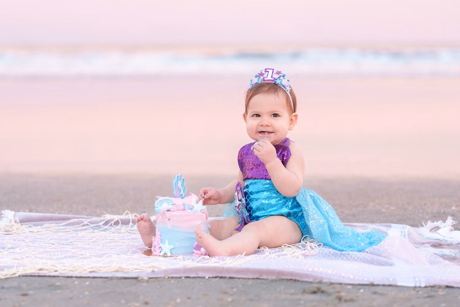 Baby girl in a mermaid outfit and crown with her cake on a blanket by the ocean in North Myrtle Beach