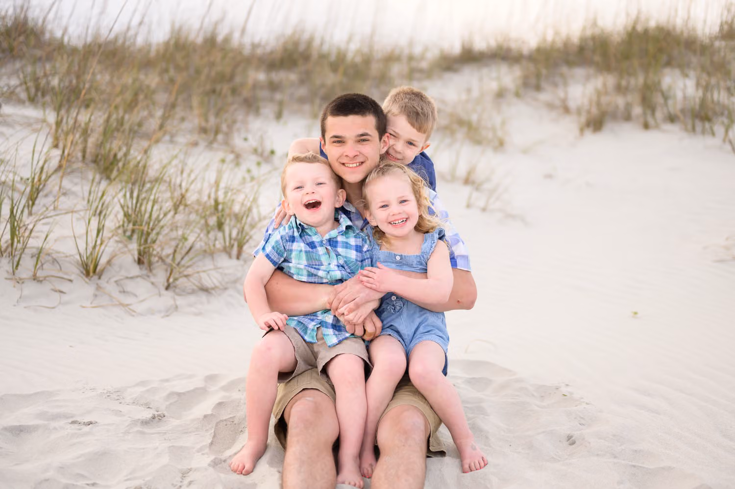 Big brother sitting in the sand by the dunes with two little kids hugging him from both sides in Pawleys Island