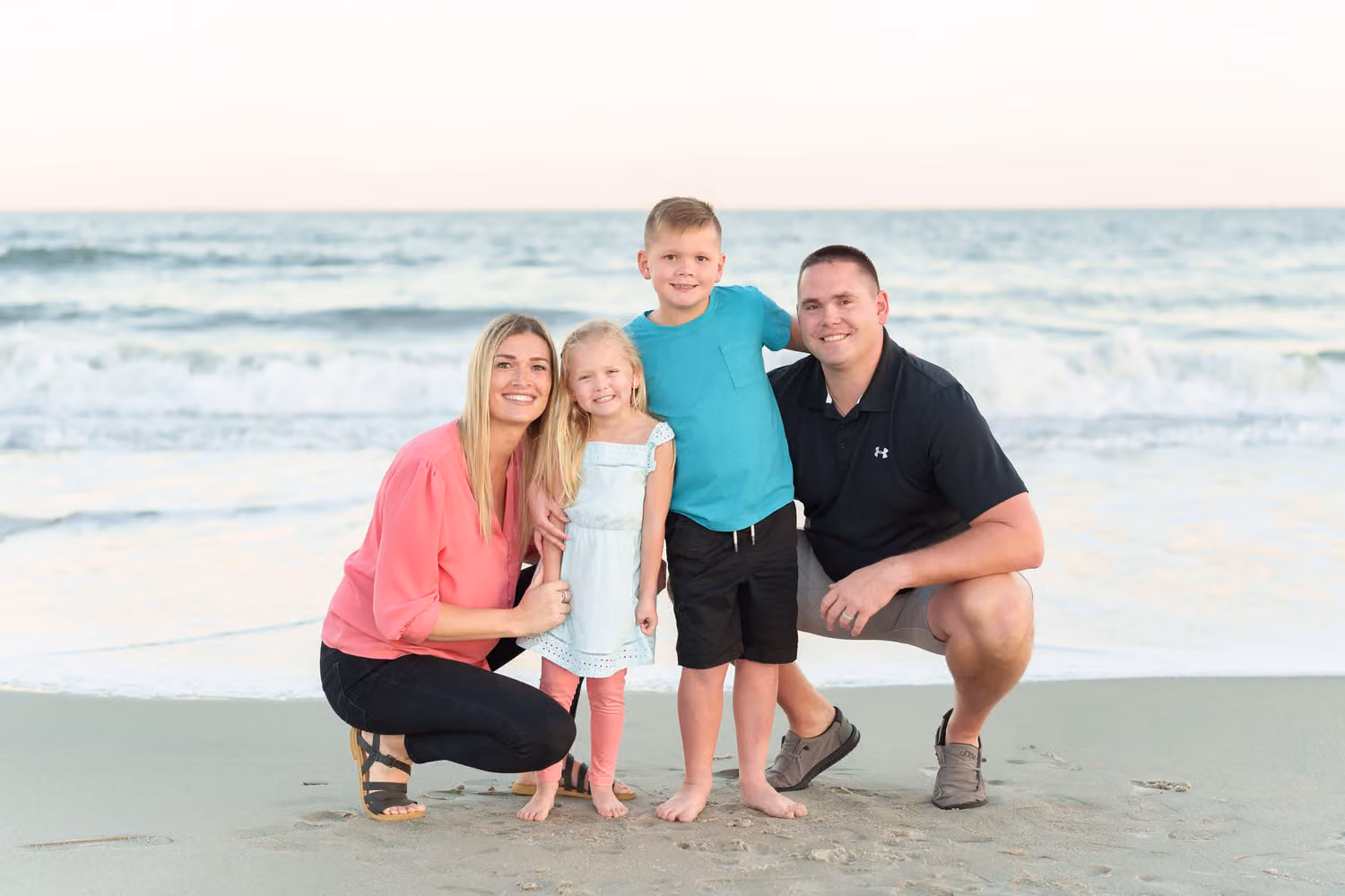 Mom and dad crouching in the sand with their son and daughter between them by the ocean at Huntington Beach State Park
