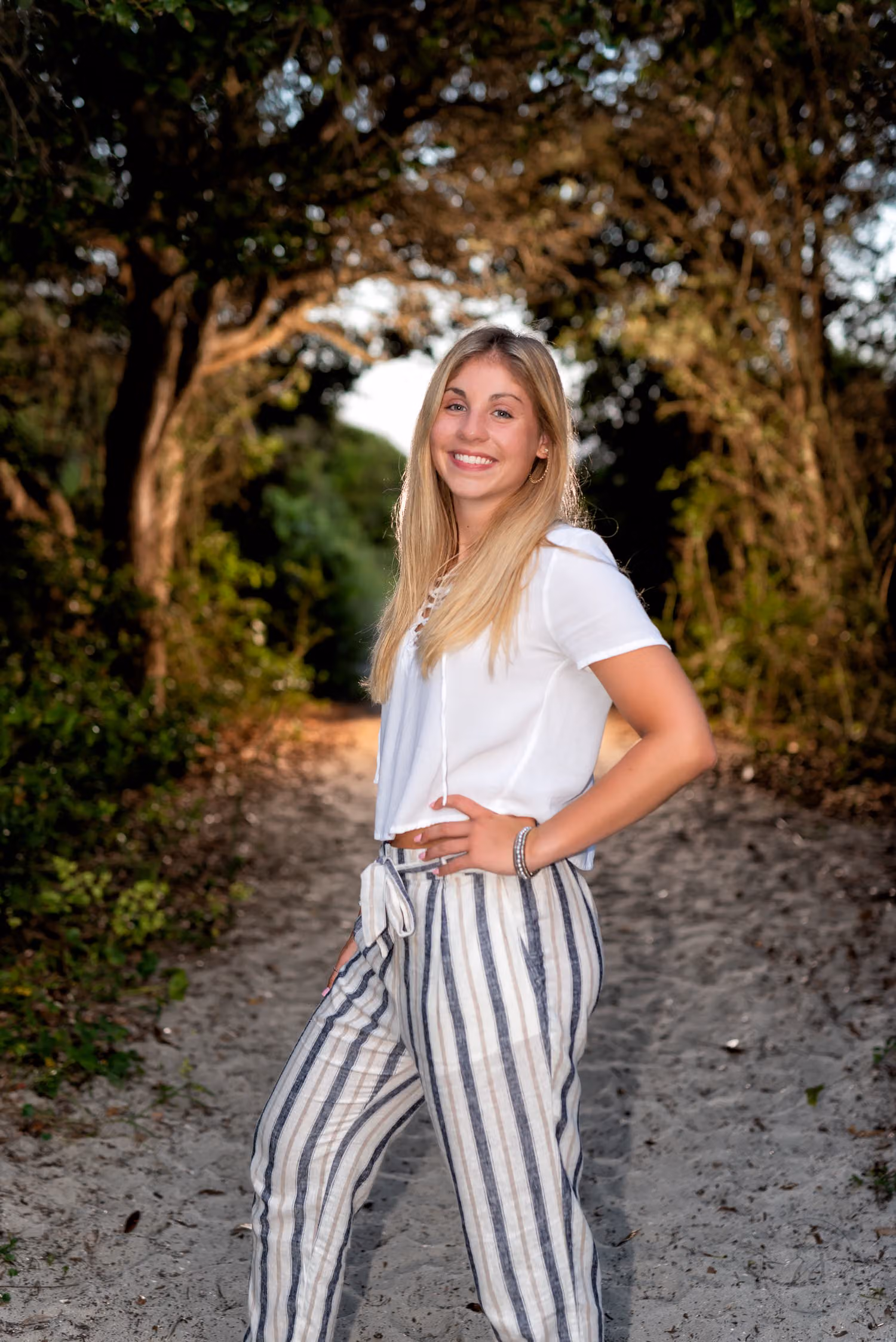 Senior in striped pants on a shaded path under an arch of trees, rim lit by flash at Huntington Beach State Park