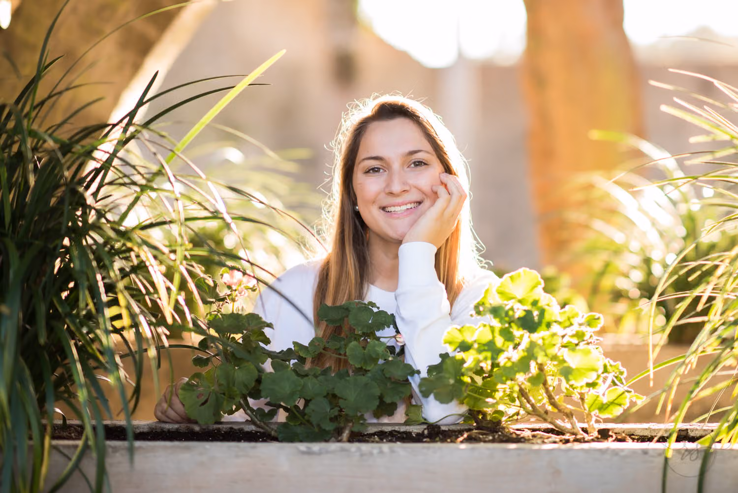 Smiling senior with her chin on her hand behind a planter, backlit by golden sunset at Atalaya Castle