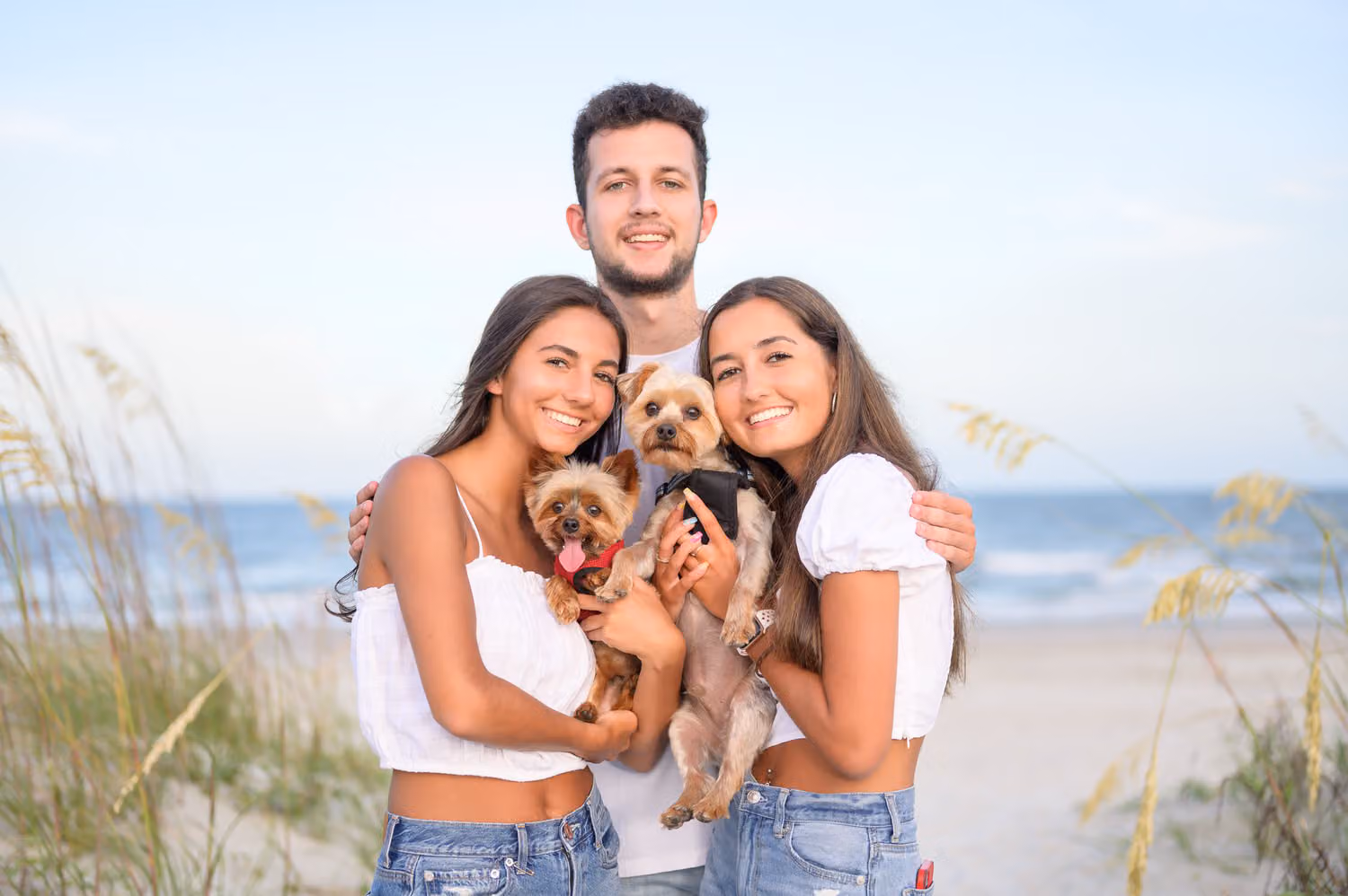 Brother standing behind his two sisters as they hold two small dogs by the sea oats at Huntington Beach State Park