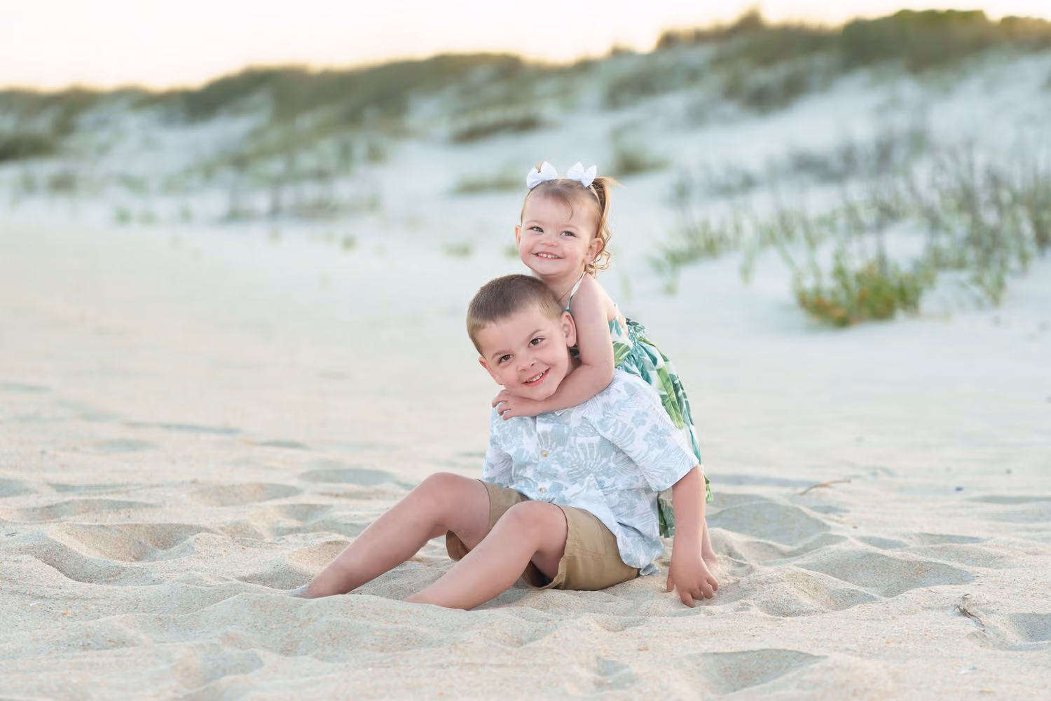 Little girl hugging her brother from behind as they sit in the sand by the dunes at Huntington Beach State Park