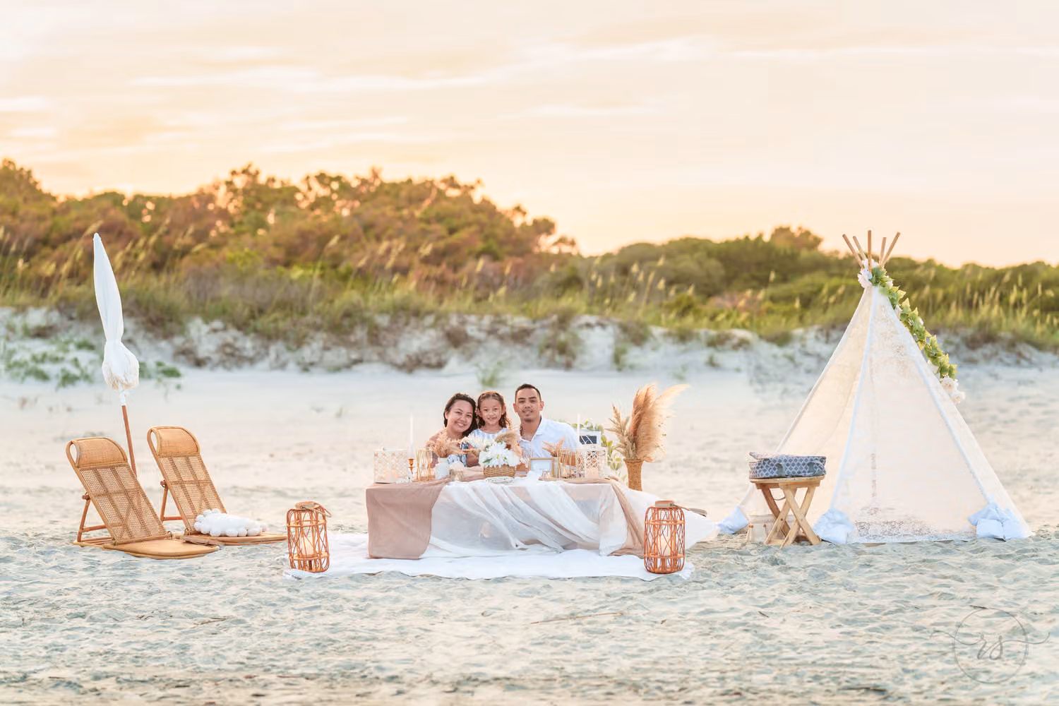 Couple and their daughter at a beach picnic table with chairs, a white teepee and dunes behind at Huntington Beach State Park