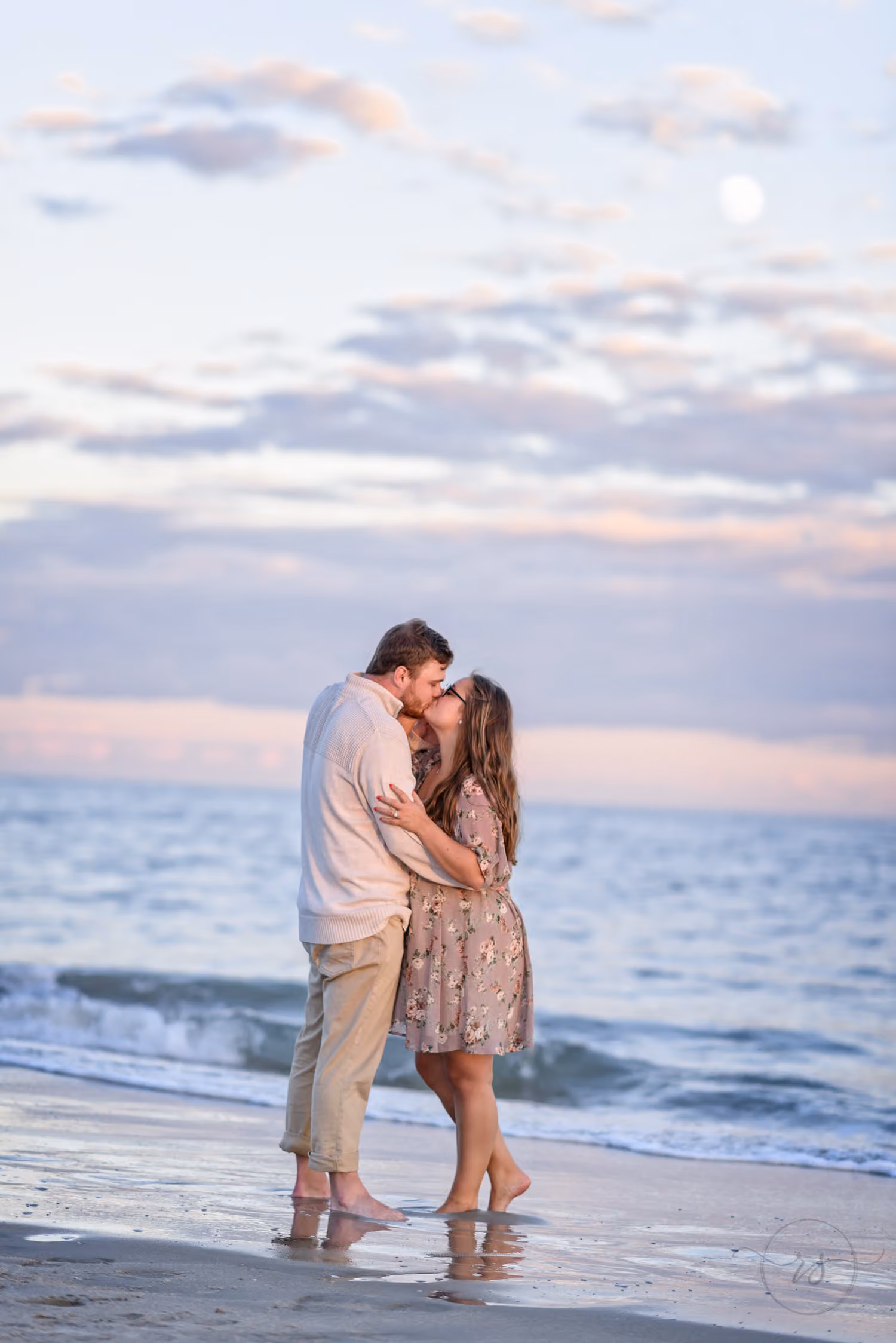 Couple kissing at the water's edge under pastel sunset clouds with the moon above at Huntington Beach State Park