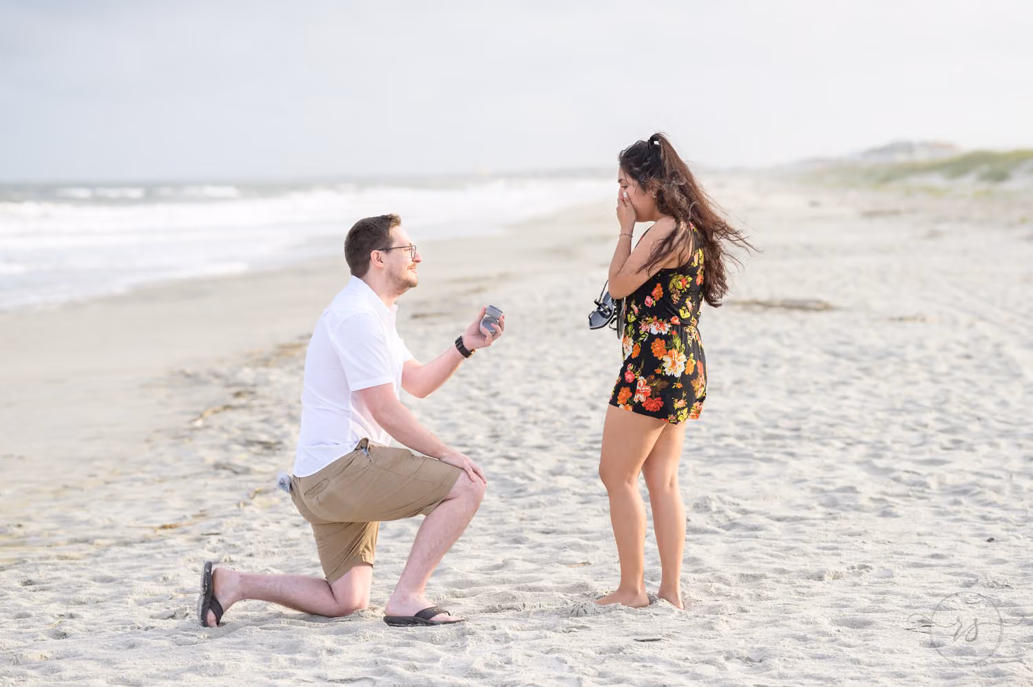 Man on one knee holding out a ring as she covers her face in surprise on the beach at Huntington Beach State Park