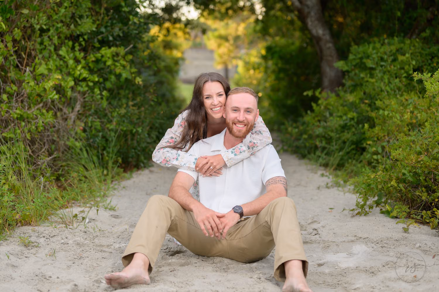 Woman hugging her fiancé from behind as they sit barefoot on a sandy path lined with brush at Huntington Beach State Park