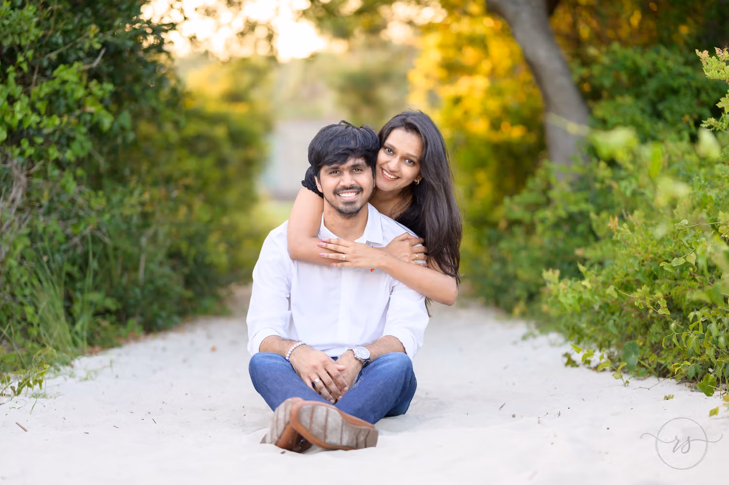 Woman hugging her fiancé from behind as they sit on a sandy path framed by green brush at Huntington Beach State Park