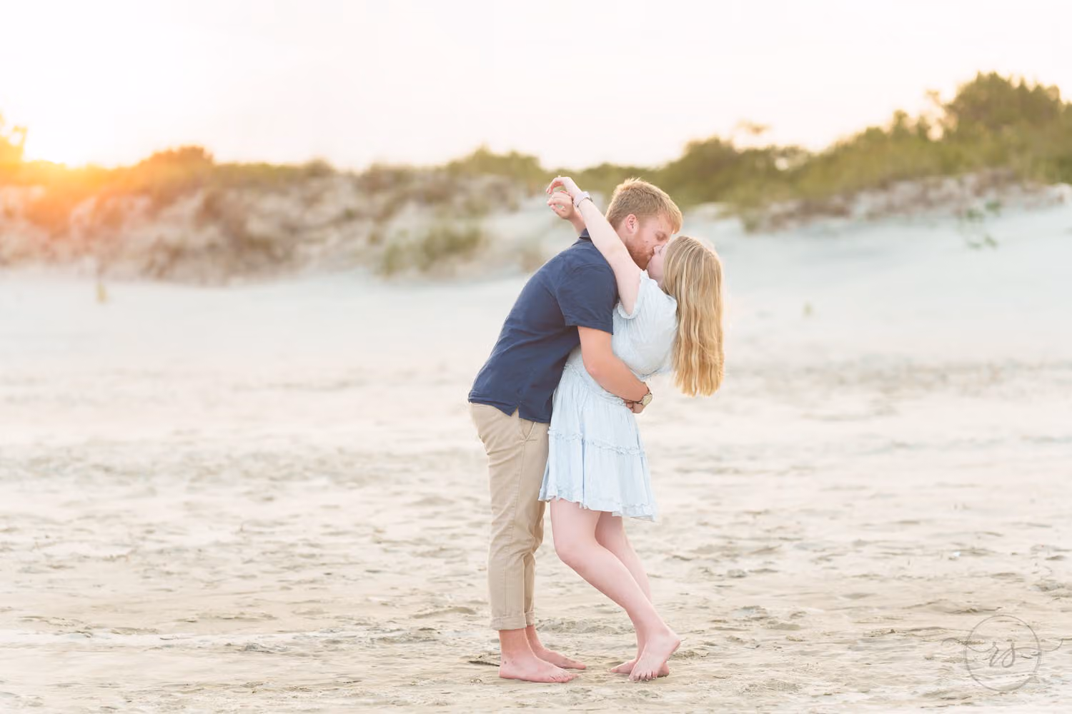 Barefoot couple sharing a kiss on the sand with golden light over the dunes at Huntington Beach State Park