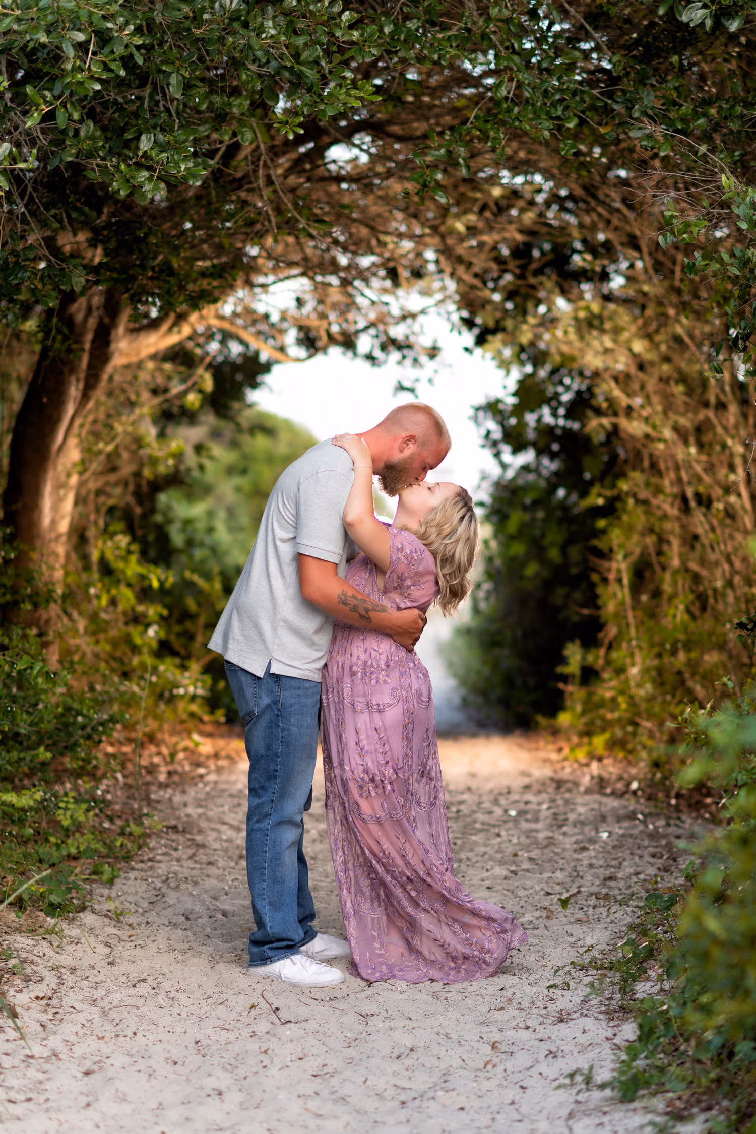 Couple kissing under an arch of trees on a sandy beach walkway, backlit by flash