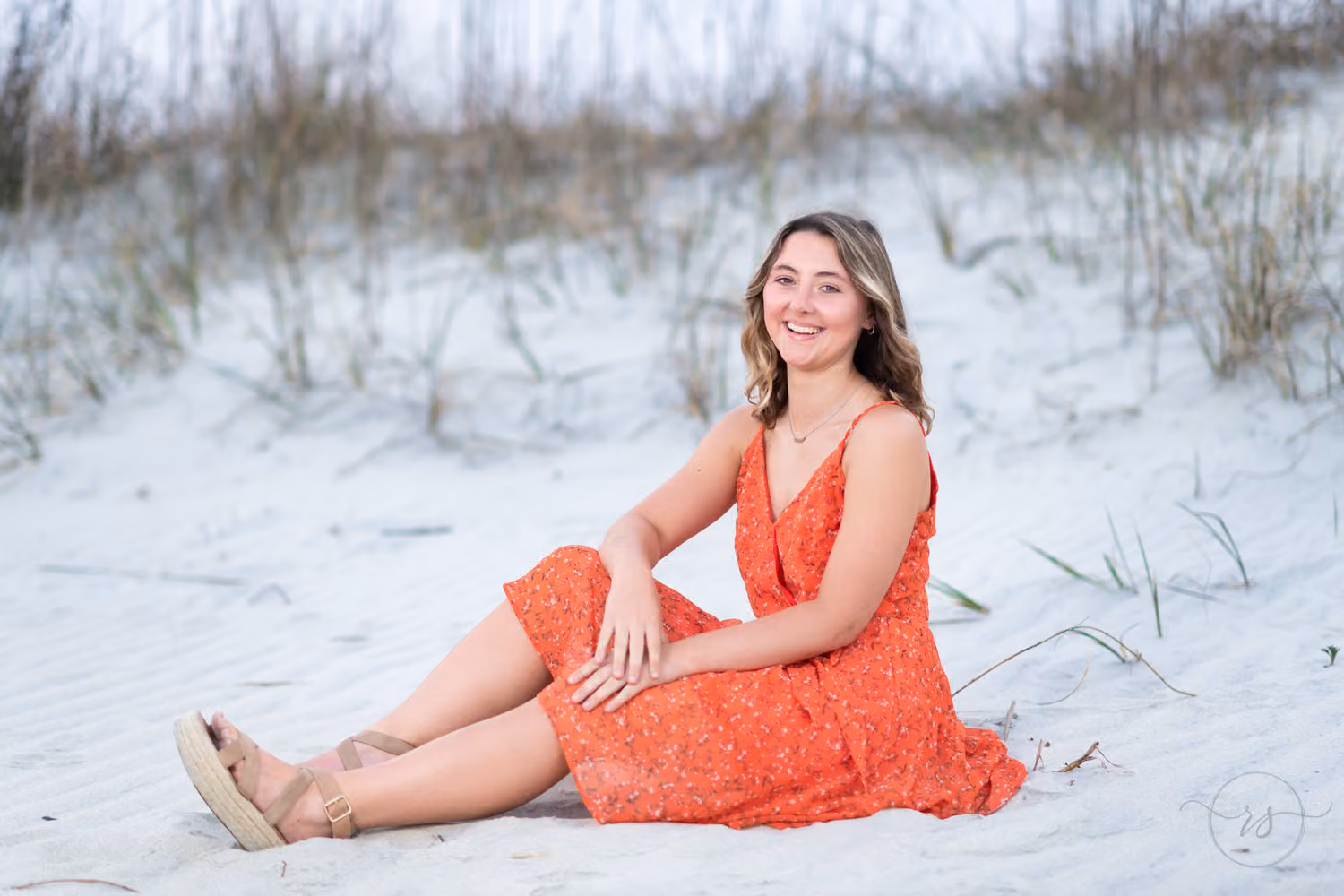 Senior in an orange lace dress sitting in the sand by the sea oats at Huntington Beach State Park
