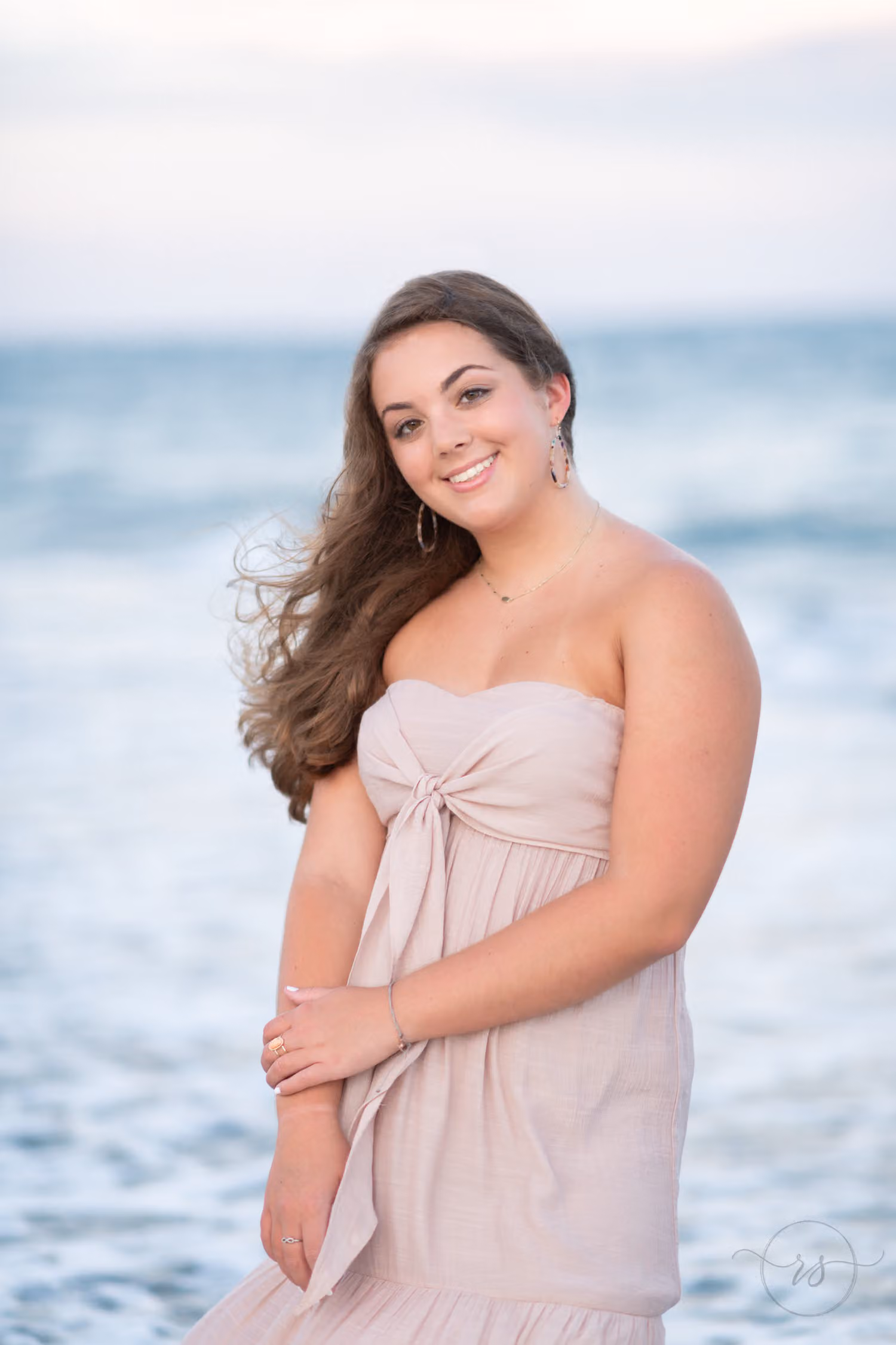 Senior in a blush strapless dress smiling with the ocean blurred behind her at Huntington Beach State Park