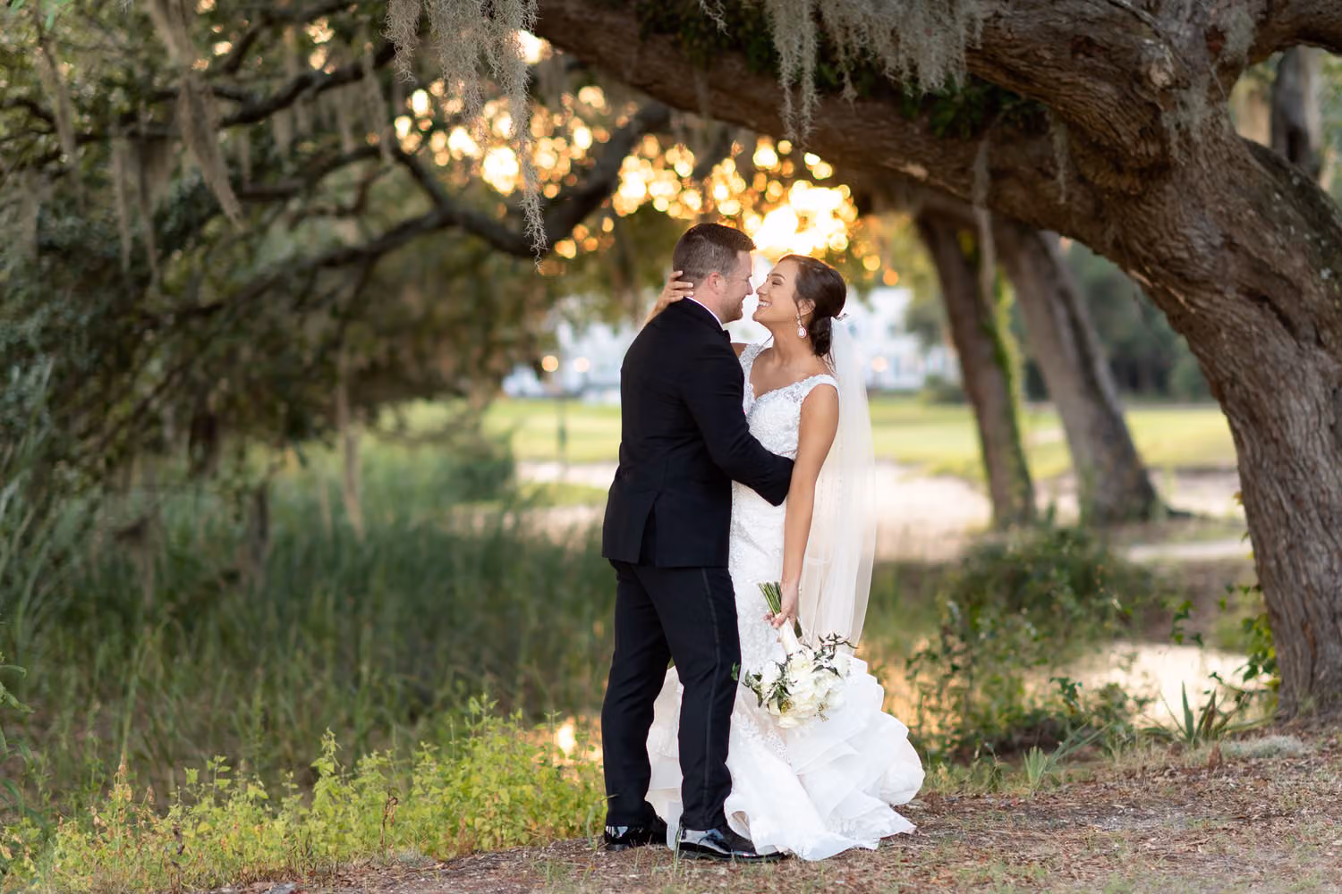 Bride and groom smiling at each other under a sweeping oak limb at the edge of the marsh at Pawleys Plantation