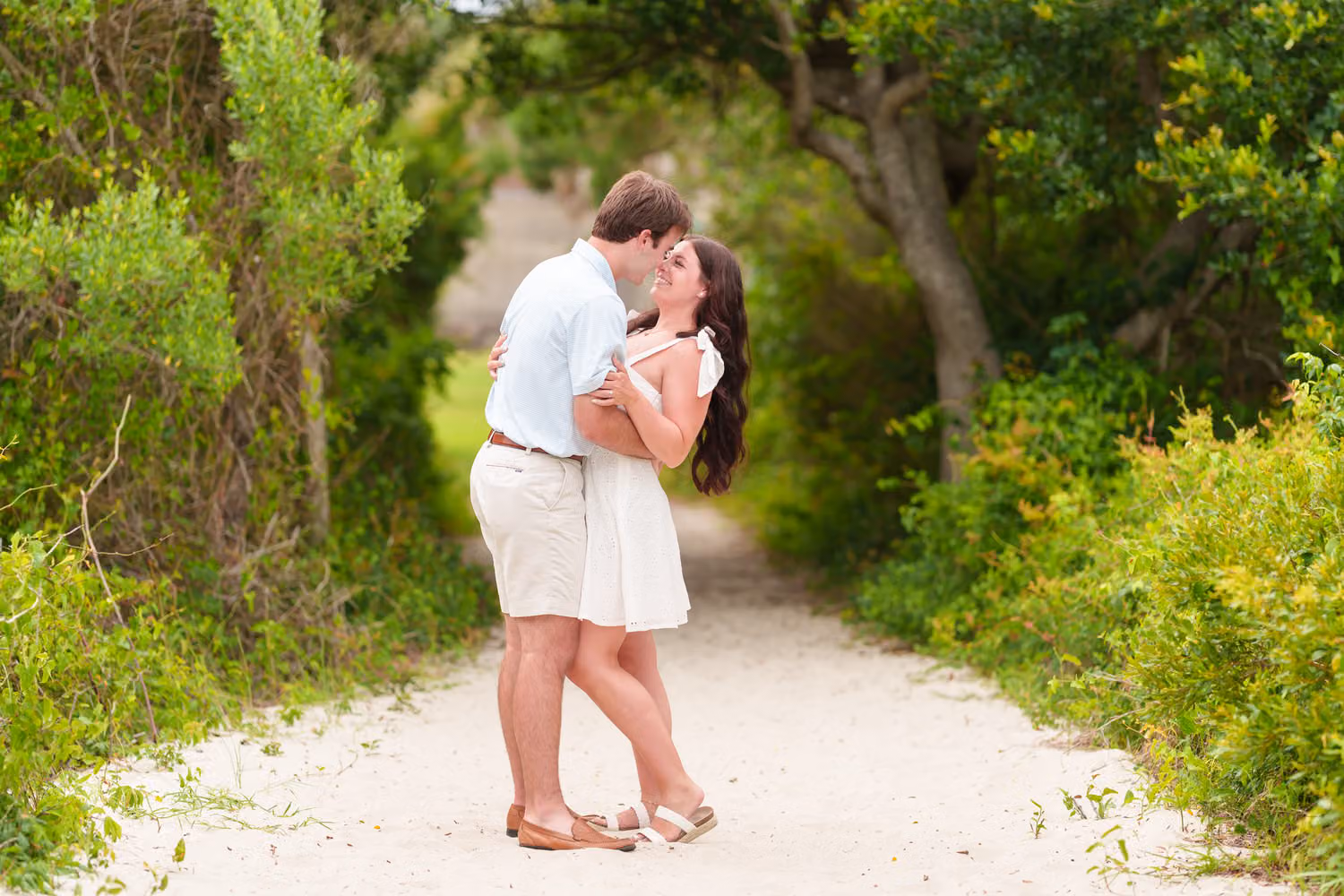 Couple kissing on a sandy path surrounded by green trees and bushes at Huntington Beach State Park