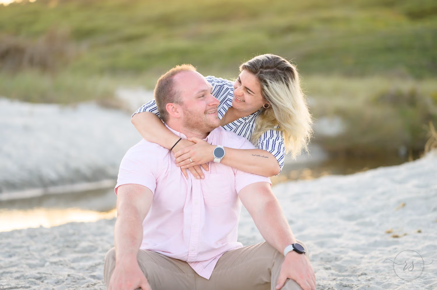 Woman hugging her fiancé from behind as they sit laughing in the sand in front of the dunes at Myrtle Beach State Park