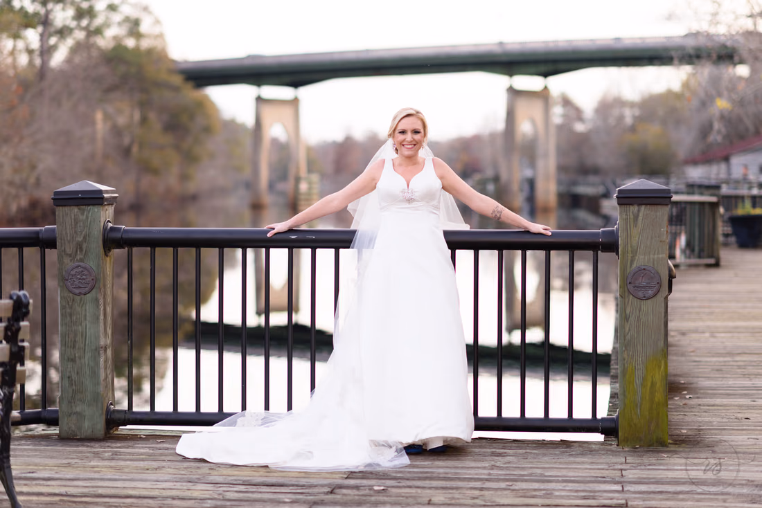 Bride with her arms spread along the boardwalk railing and the bridge behind her at the Conway River Walk