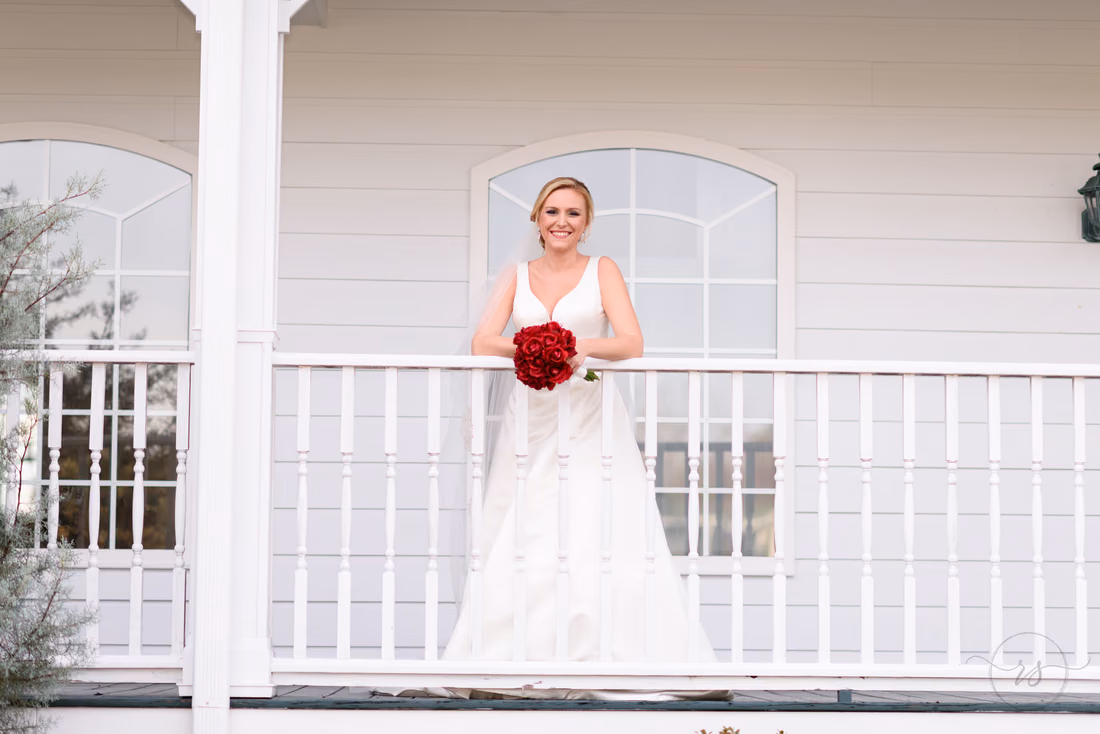 Bride leaning on a white porch railing with her red rose bouquet near the Conway River Walk