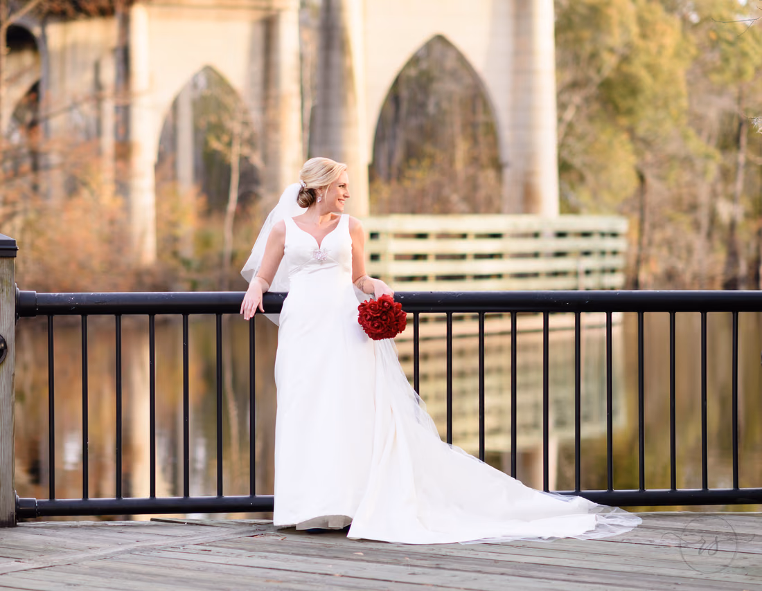 Bride leaning on the boardwalk railing with the arches of the old bridge behind her at the Conway River Walk