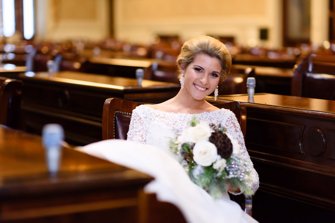 Smiling bride holding her bouquet at a wooden desk in a legislative chamber of the South Carolina State House