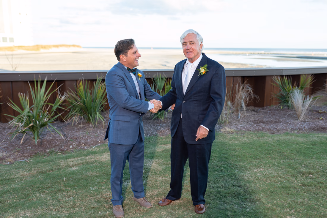 Groom shakes hands with his son and best man on the lawn by the ocean at the Dunes Golf and Beach Club in Myrtle Beach