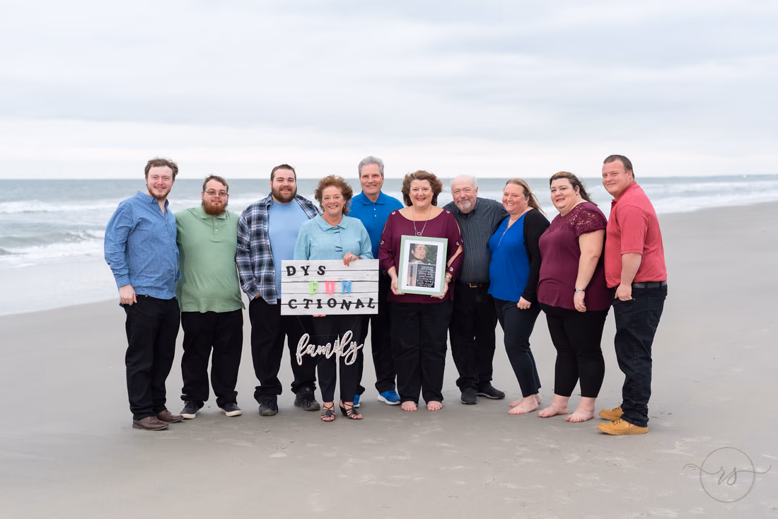 Large family on the beach holding a letter board sign and a framed photo at Huntington Beach State Park