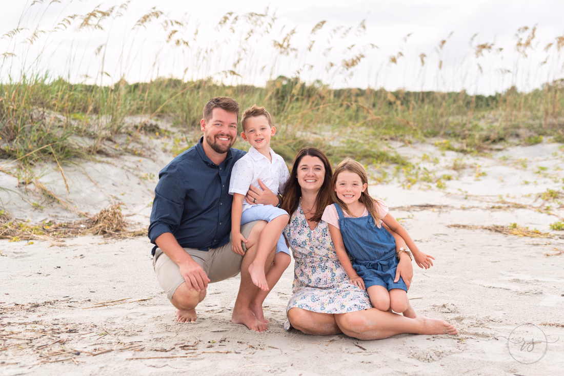 Mom, dad and their two kids sitting in the sand in front of the sea oats at Huntington Beach State Park