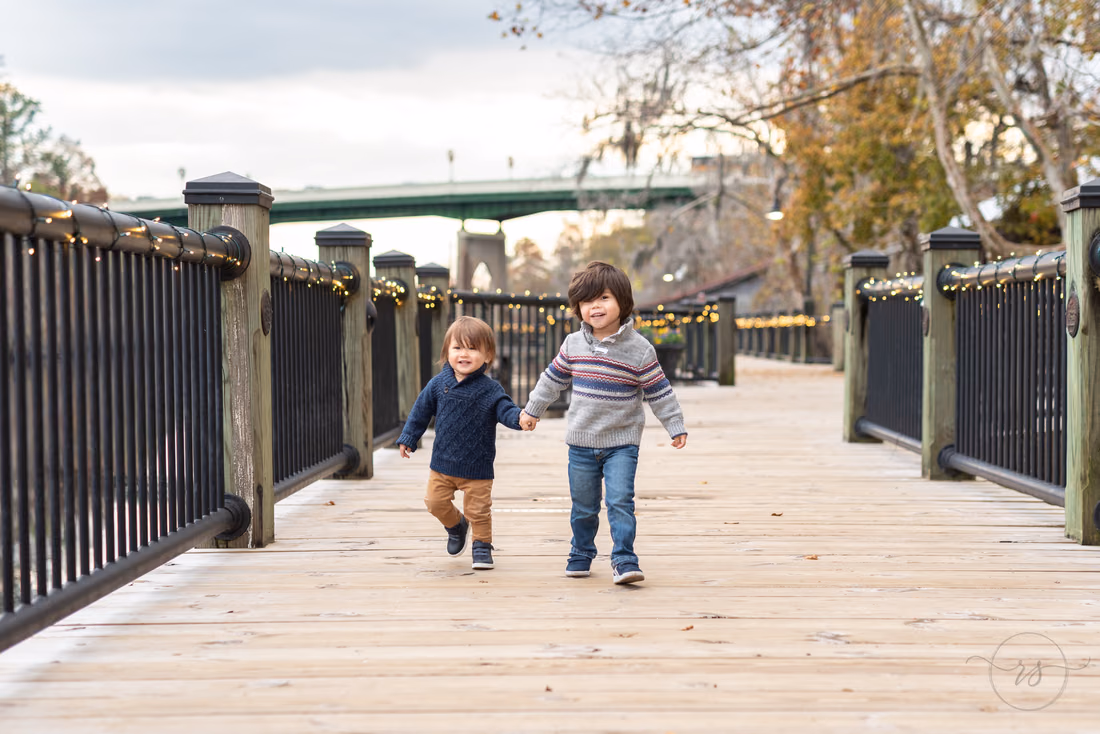 Two little boys walk hand in hand down the boardwalk with the bridge behind them at the Conway Riverwalk
