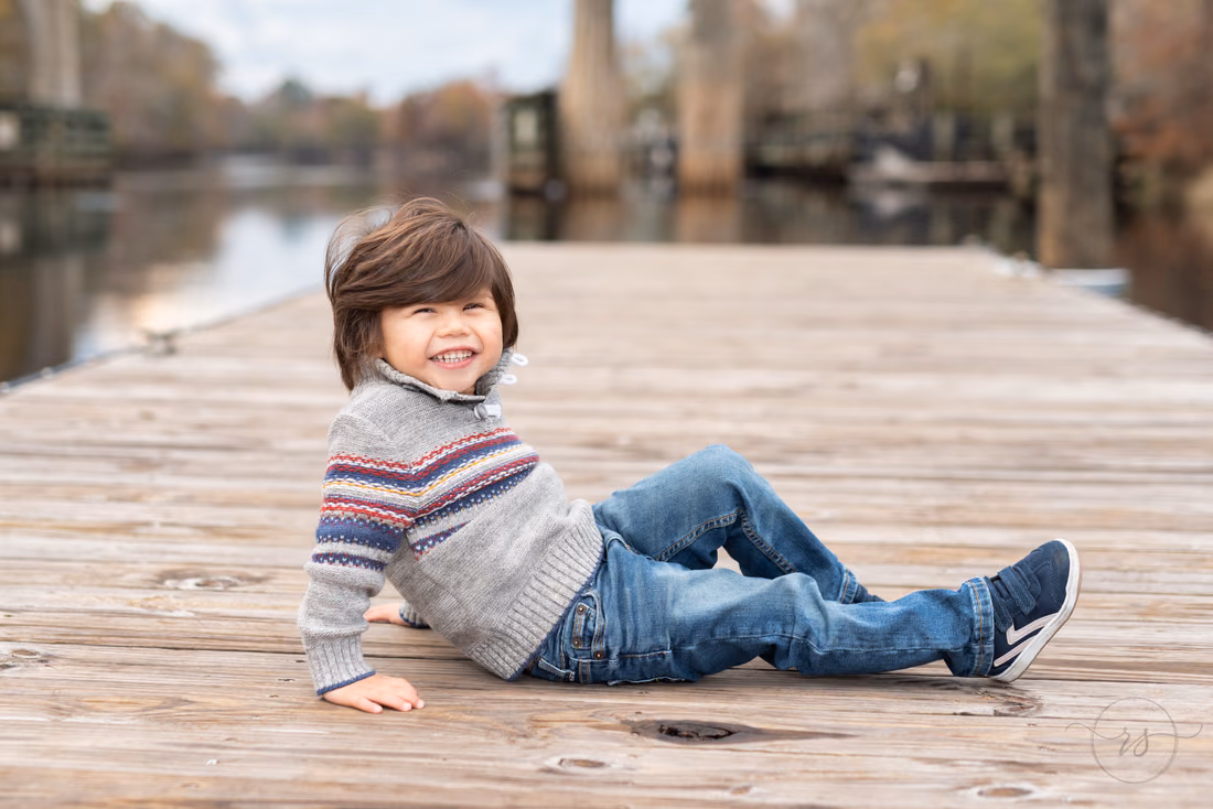 Little boy in a striped sweater laughs as he leans back on the dock by the river at the Conway Riverwalk