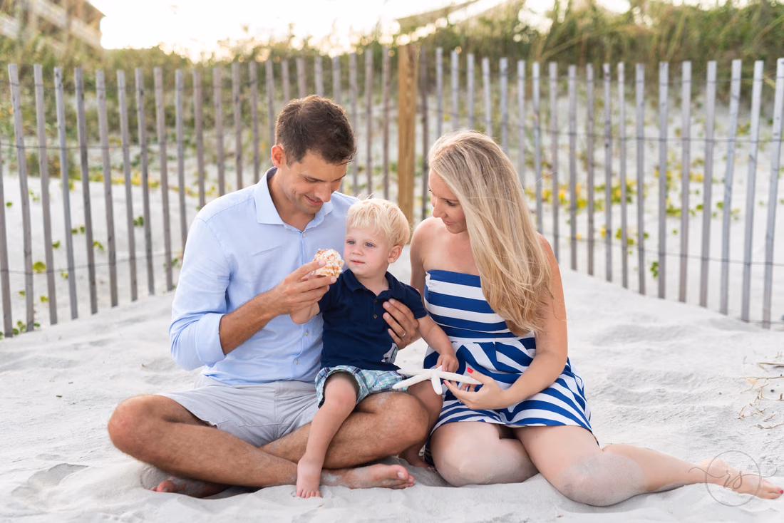Mom, dad and their toddler sitting in the sand by a dune fence on the beach
