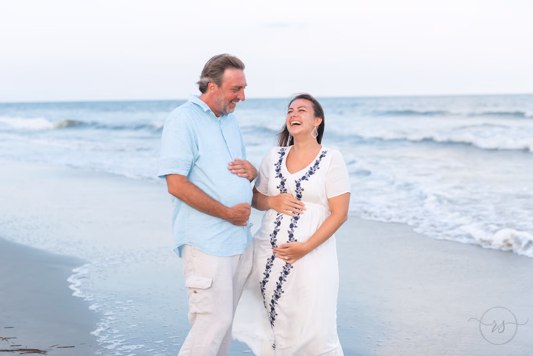 Expecting couple laughing together at the water's edge at Litchfield Beach