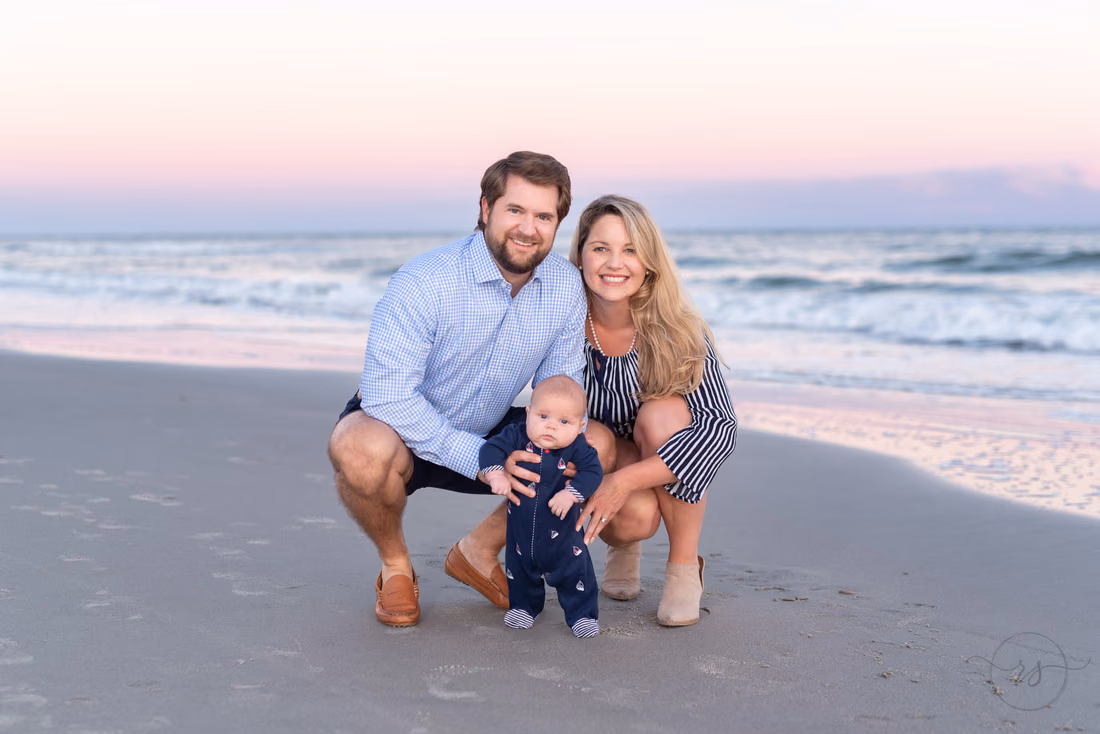 Parents crouch on the beach at sunset helping their baby boy stand at Huntington Beach State Park