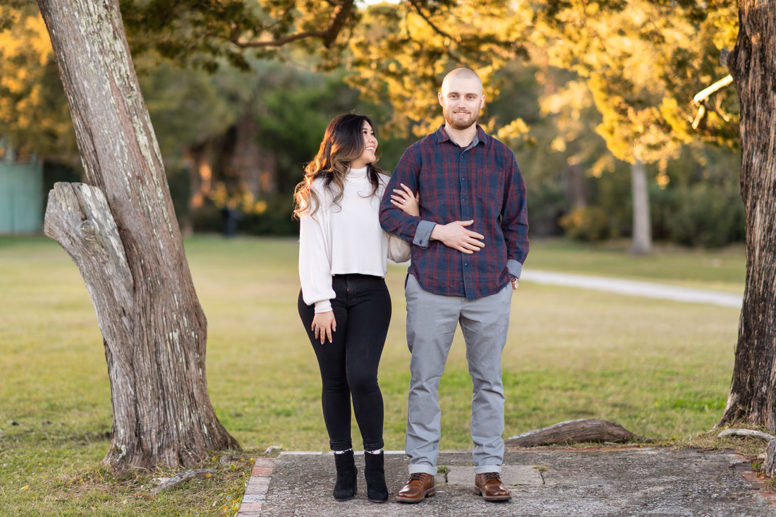 Couple stand arm in arm between two old oak trunks at Huntington Beach State Park