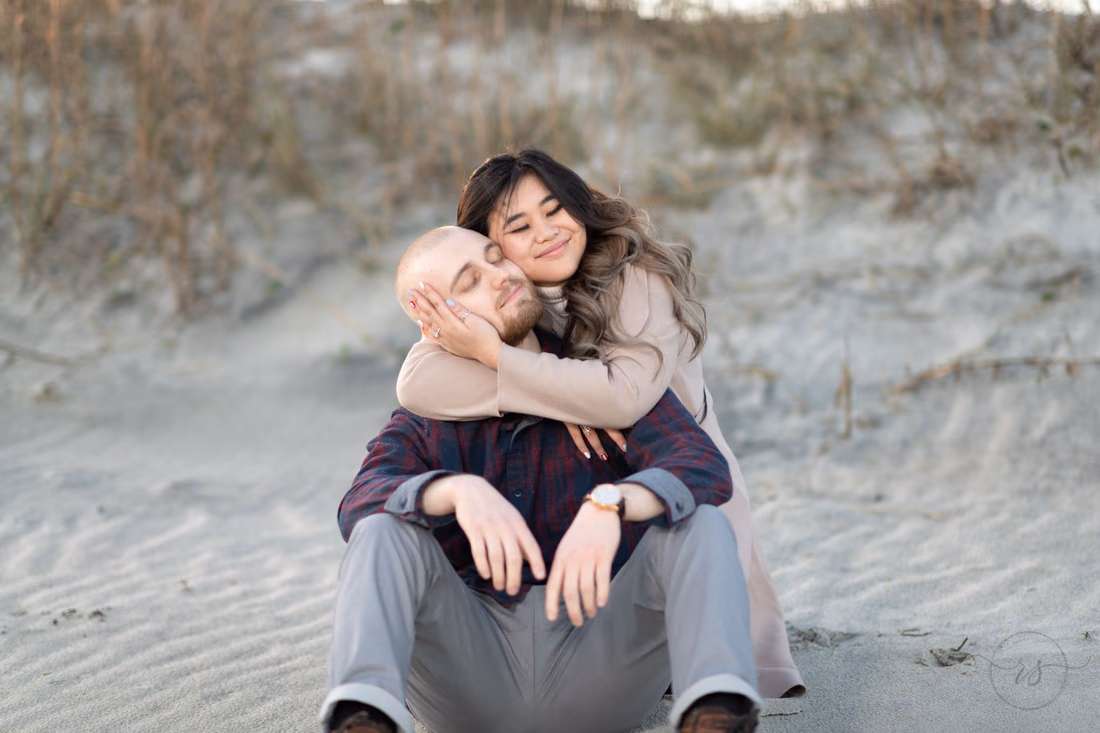 She hugs him from behind as they sit in the sand in front of the dunes at Huntington Beach State Park