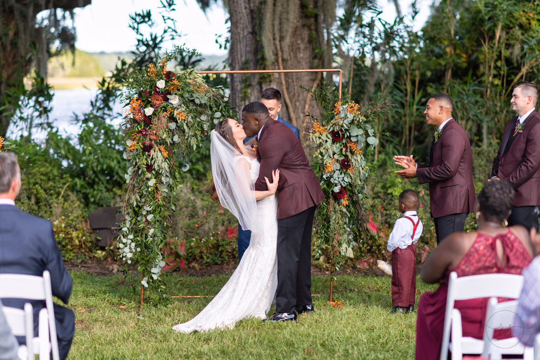 Bride and groom kiss under the floral arch as guests applaud at Magnolia Plantation