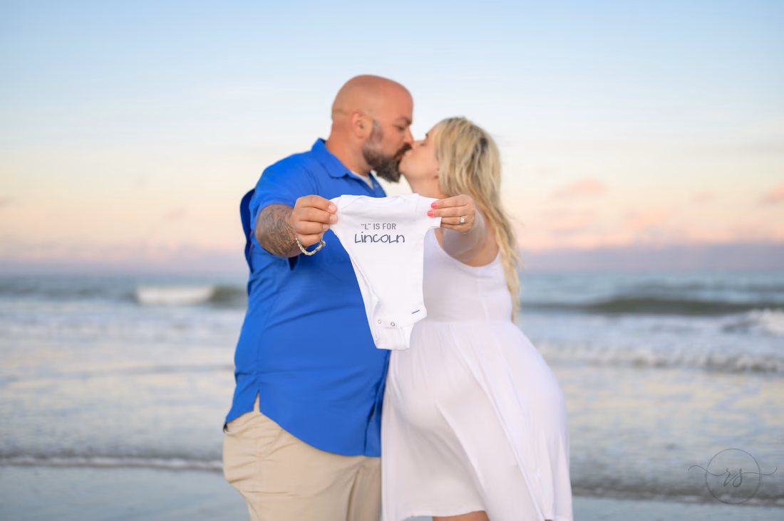 Couple kisses while holding up a baby onesie with the baby's name at Huntington Beach State Park