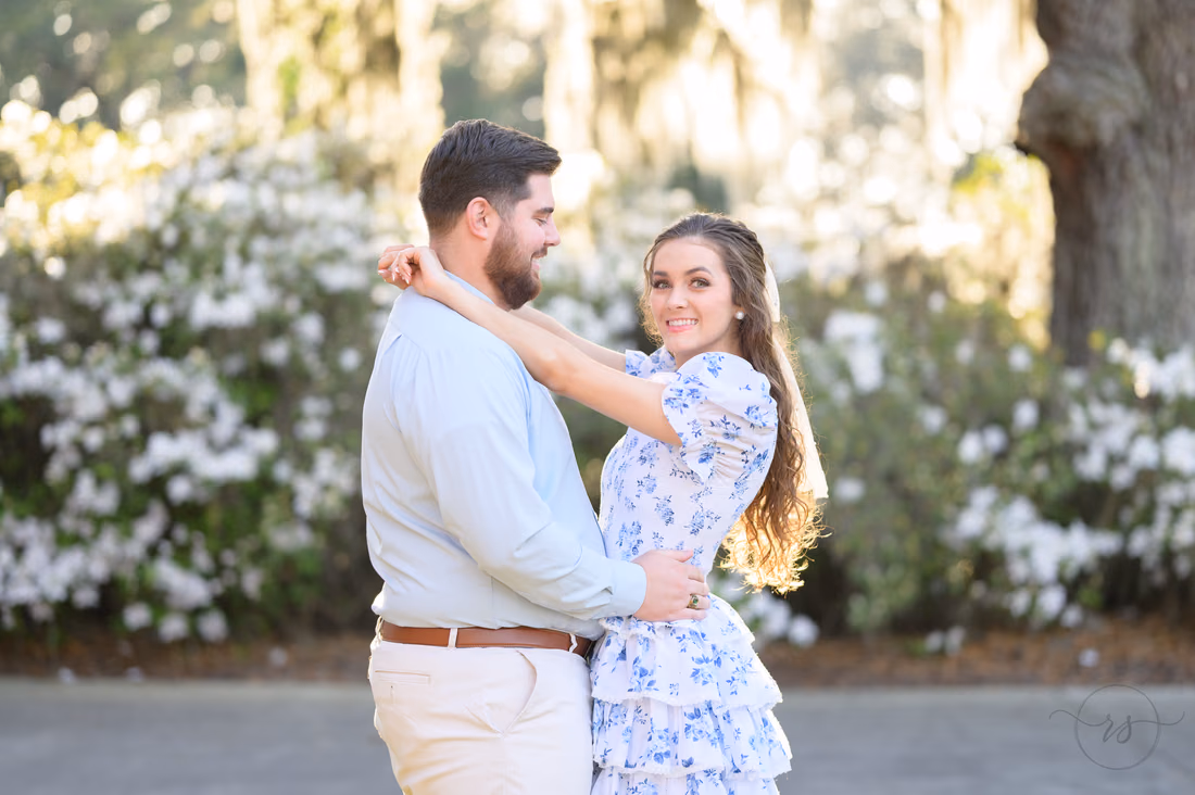 Engaged couple embracing in front of white azaleas at Caledonia Golf and Fish Club