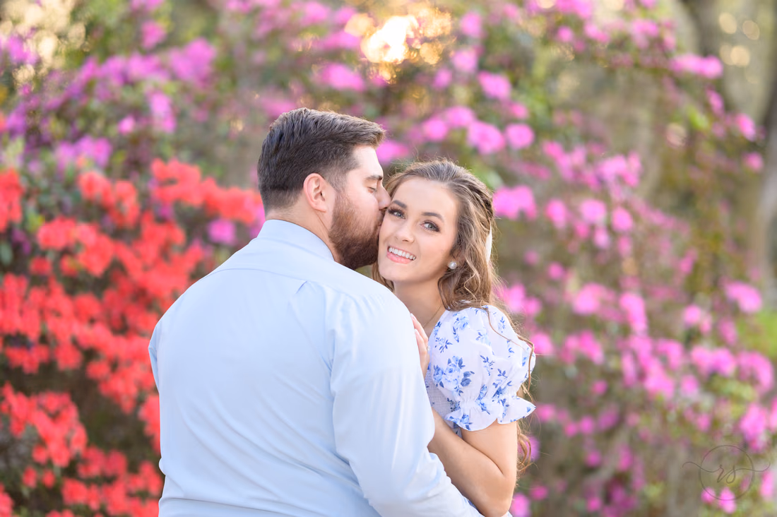 He kisses her cheek in front of red and purple azaleas at Caledonia Golf and Fish Club