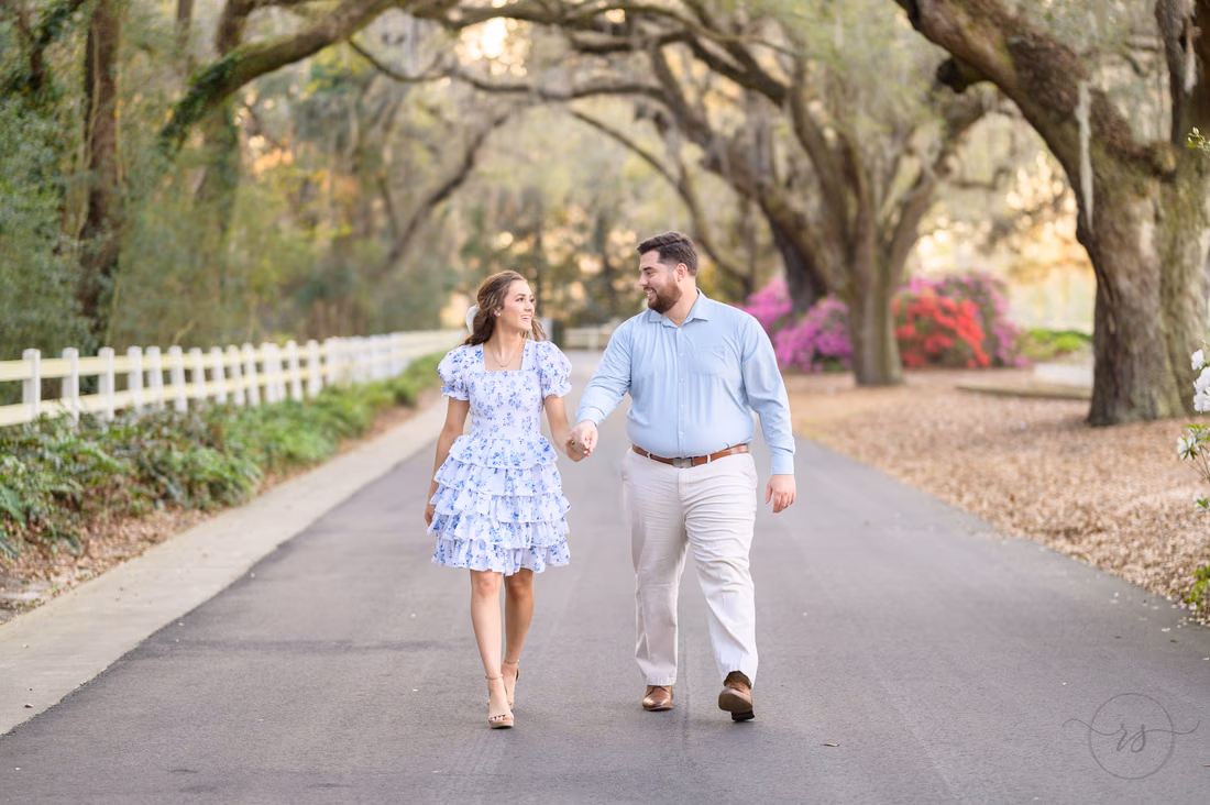 Couple walking hand in hand toward the camera under the oak-lined road at Caledonia Golf and Fish Club