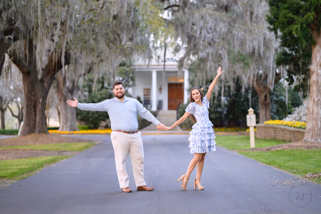 Couple holding hands with arms stretched wide in front of the white clubhouse at Caledonia Golf and Fish Club