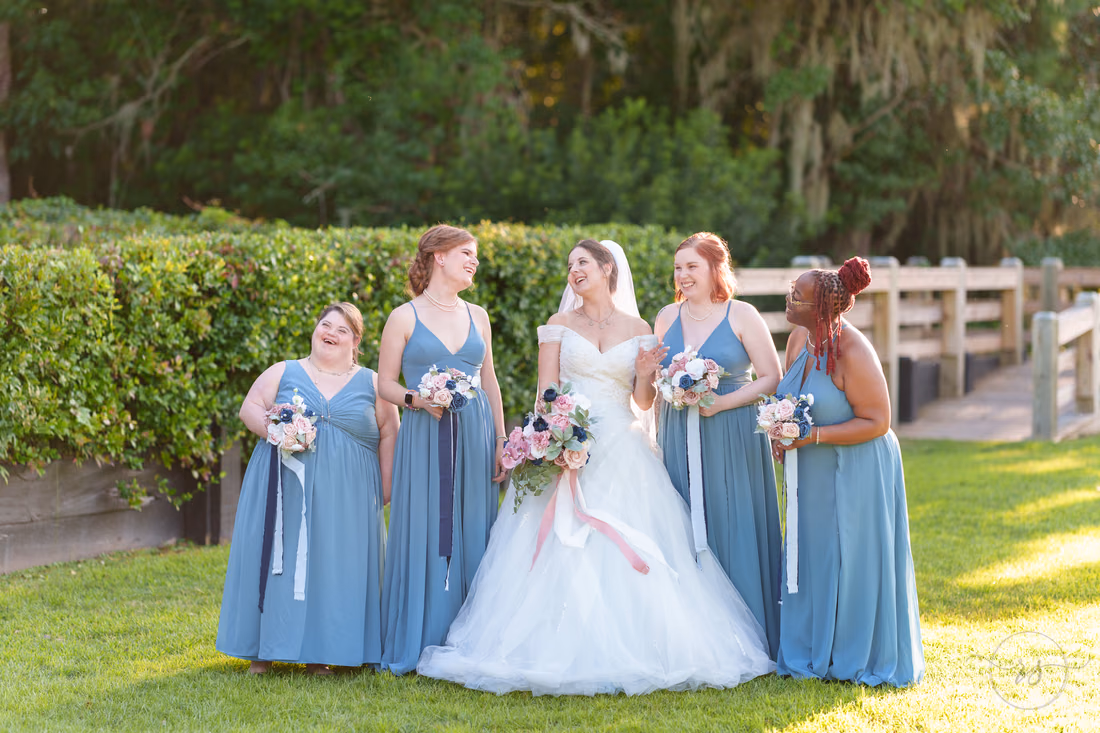 Bride and bridesmaids in dusty blue laughing together by the hedge - The Pavilion at Pepper Plantation