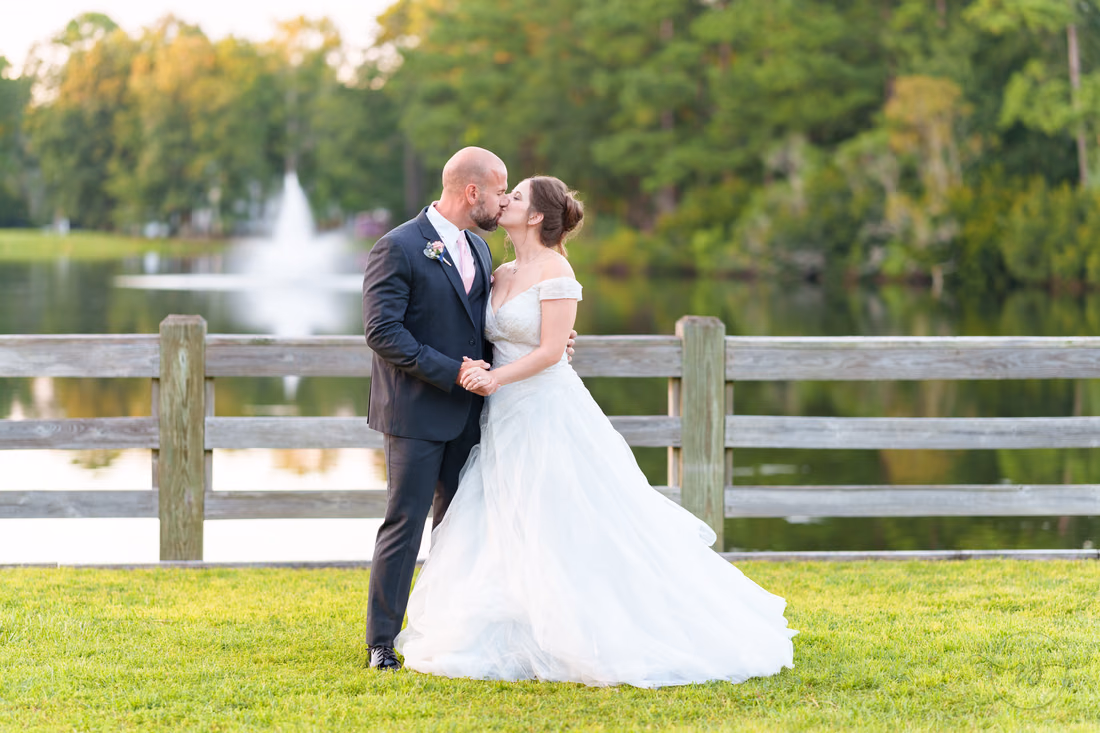Bride and groom kissing by the fence with the lake fountain behind them - The Pavilion at Pepper Plantation