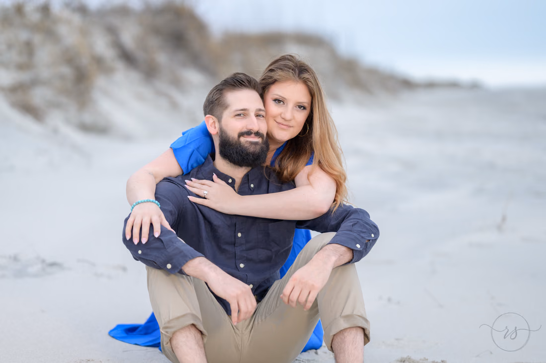 She hugs him from behind as they sit in front of the dunes - Huntington Beach State Park