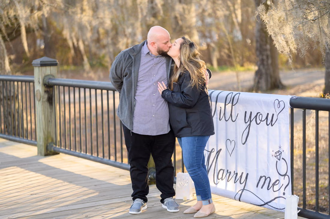 Couple kissing beside a Will you marry me banner on the boardwalk - Conway River Walk