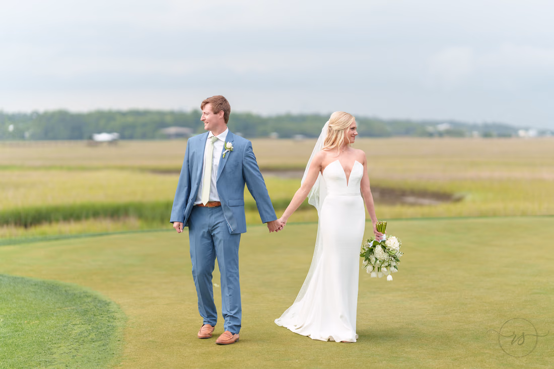 Bride and groom walk hand in hand across the green by the marsh at Pawleys Plantation