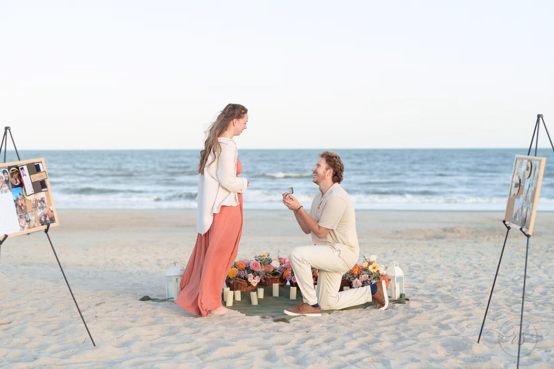 Man on one knee proposing beside flowers and photo displays on the beach - Huntington Beach State Park