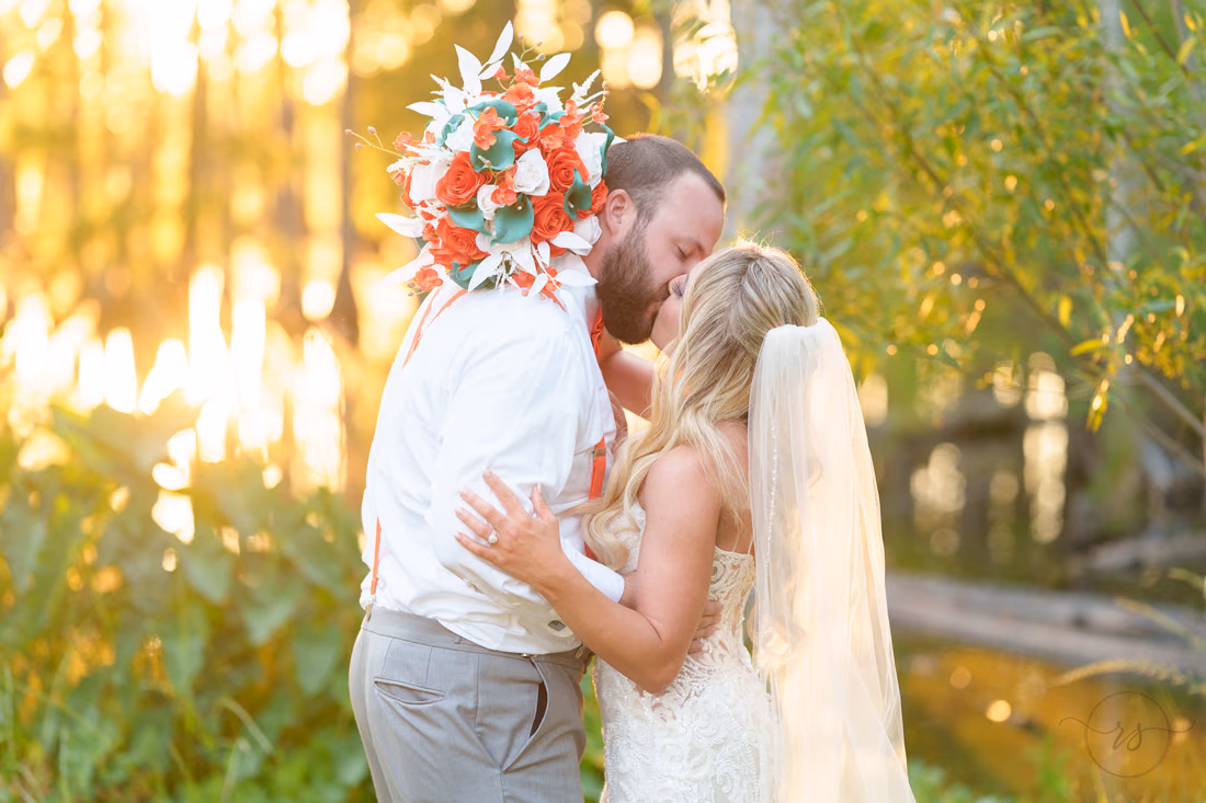 Bride and groom kissing as she holds her bouquet up in the golden light by the marsh at Wildhorse at Parker Farms